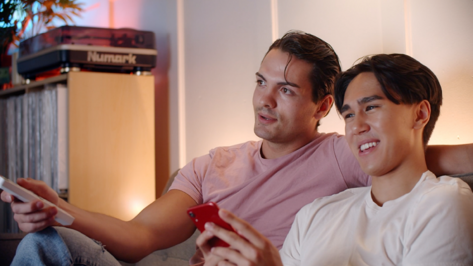 Greg and Brad sit on the sofa browsing the TV while Greg texts on his red phone.